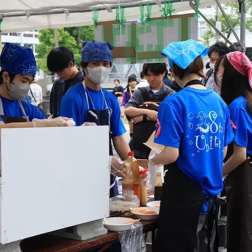 大宮祭の模擬店で、青いTシャツとバンダナを身に着けた学生スタッフがマスクと手袋を着用し、鉄板前で調理しながら来場者対応を行っている